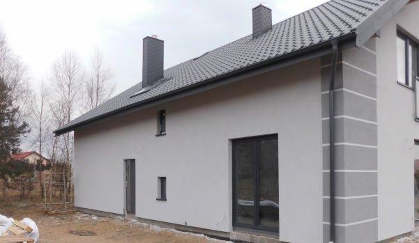 Nowoczesny dom jednorodzinny z szarą elewacją, ciemnoszarą dachówką i kominami, widoczne rynny, okna i drzwi. Na elewacji dekoracyjne pasy w odcieniu szarości. W tle drzewa bez liści i fragment...