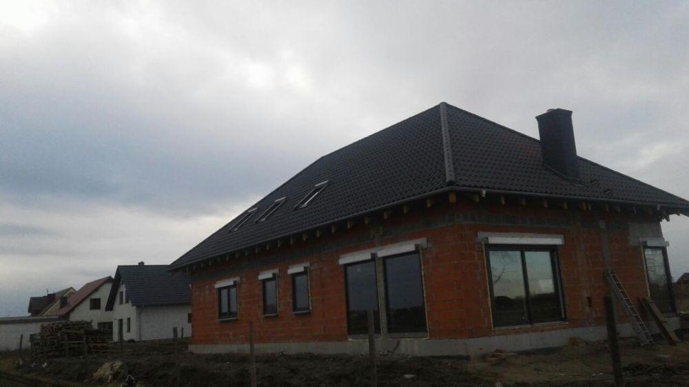 Surowy, niewykończony dom z czerwonej cegły z ciemnym dachem i kominem, widoczne okna z roletami, drabina oparta o ścianę, pochmurne niebo w tle.