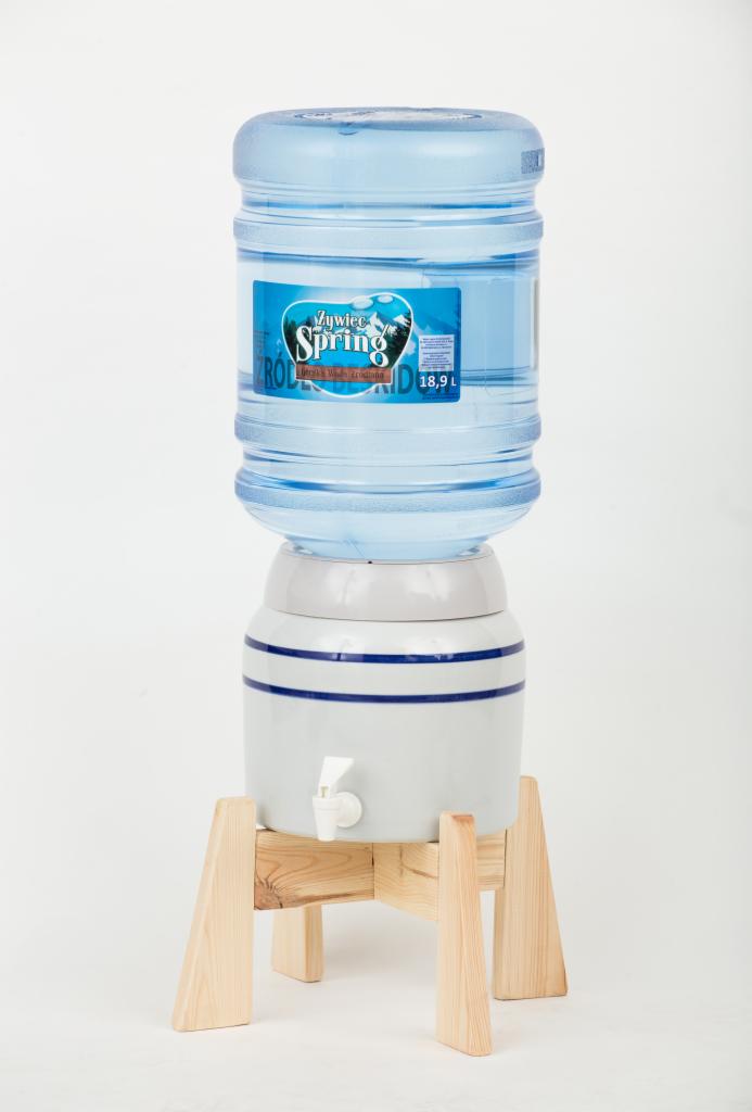 Dystrybutor wody z butlą 18,9l marki Żywiec Spring, umieszczony na ceramicznej podstawie z niebieskimi paskami i drewnianym stojaku.