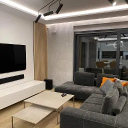 Nowoczesny salon z szarą sofą narożną, białymi meblami RTV, drewnianymi lamelami na ścianie i oświetleniem punktowym na suficie.