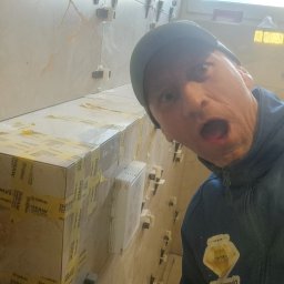 DIAMOND HOME MARCIN KOSTECKI - Mężczyzna z otwartymi ustami w bluzie i czapce na tle ściany z jasnych płytek, częściowo oklejonych taśmą, widoczne kliny poziomujące płytki.