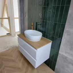 Umywalka nablatowa owalna z baterią w kolorze złotym, zamontowana na szafce z dwiema szufladami, na podłodze parkiet ułożony w jodełkę, w tle ściana wyłożona wąskimi, zielonymi płytkami.