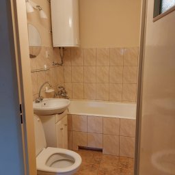 DIAMOND HOME MARCIN KOSTECKI - Ustawna łazienka w beżowych tonacjach z wanną obudowaną płytkami, umywalką z szafką i toaletą. Na ścianie bojler i lustro z oświetleniem. Widok zza uchylonych drzwi.