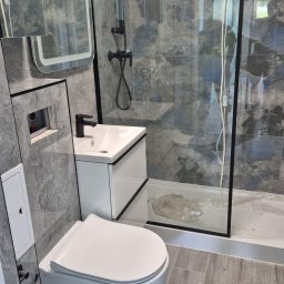 DIAMOND HOME MARCIN KOSTECKI - Nowoczesna łazienka po remoncie: wisząca toaleta, umywalka z szafką, kabina prysznicowa z czarnymi ramami, szare płytki imitujące kamień i drewnopodobna podłoga.