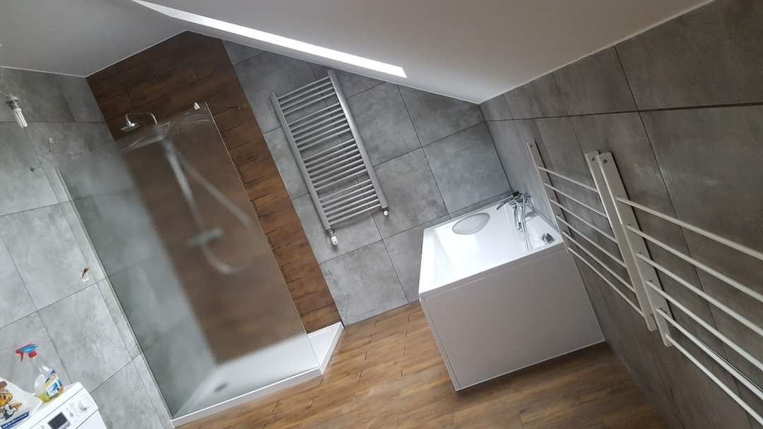 Nowoczesna łazienka w Świnoujściu z kabiną prysznicową typu walk-in, grzejnikiem drabinkowym w kolorze srebrnym, białą wanną i drewnopodobną podłogą oraz szarą płytką na ścianach.