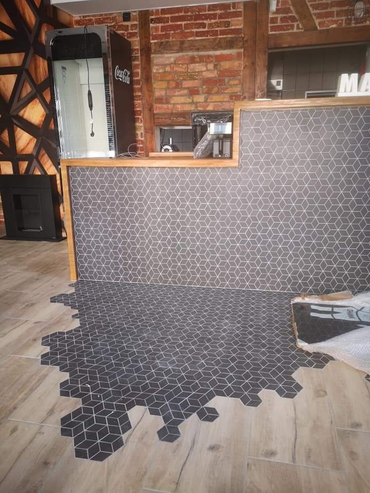 Układanie heksagonalnych płytek podłogowych w lokalu, z widocznym fragmentem ściany wykończonej szarą mozaiką w geometryczne wzory oraz niezakończony obszar łączenia z drewnopodobną podłogą.