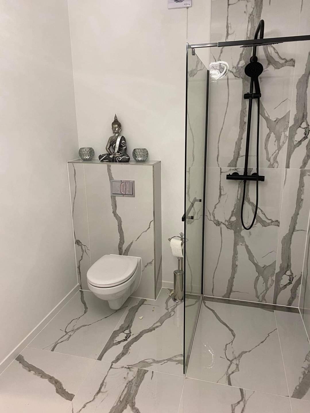 Nowoczesna łazienka z toaletą podwieszaną, kabiną prysznicową typu walk-in z czarną armaturą i płytkami imitującymi marmur, uzupełniona figurką Buddy na obudowie stelaża.