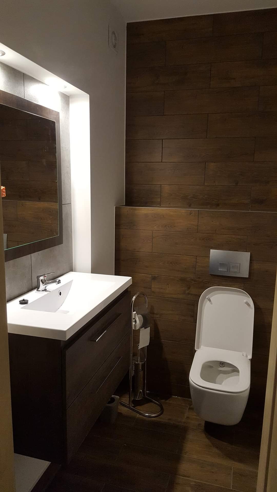 Nowoczesna łazienka z drewnopodobnymi płytkami na ścianie, wisząca toaleta z chromowanymi przyciskami spłukiwania, umywalka nablatowa na ciemnej szafce, lustro z podświetleniem LED i chromowany...