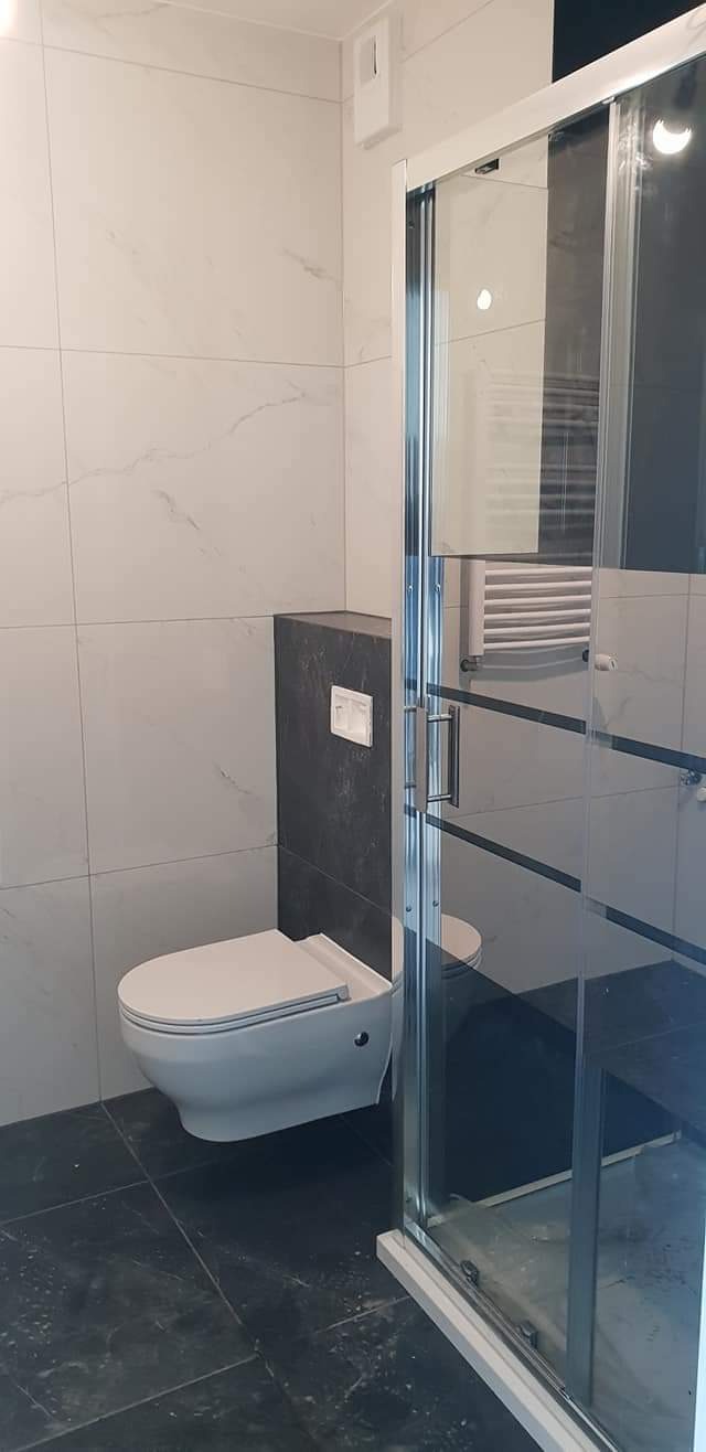 Nowoczesna łazienka z podwieszaną toaletą w zabudowie z ciemnoszarego kamienia, kabiną prysznicową ze szklanymi drzwiami przesuwnymi i białymi płytkami imitującymi marmur na ścianach.