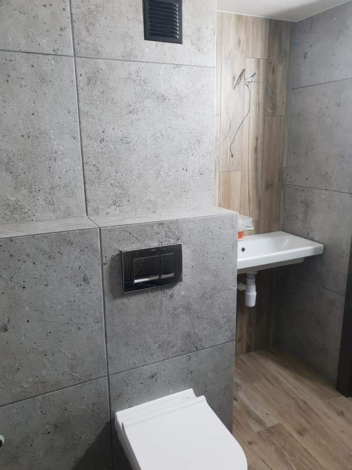 Wykończona łazienka z szarymi płytkami imitującymi beton, drewnopodobną ścianą, podwieszaną umywalką i wiszącą toaletą z czarnym przyciskiem spłukiwania.