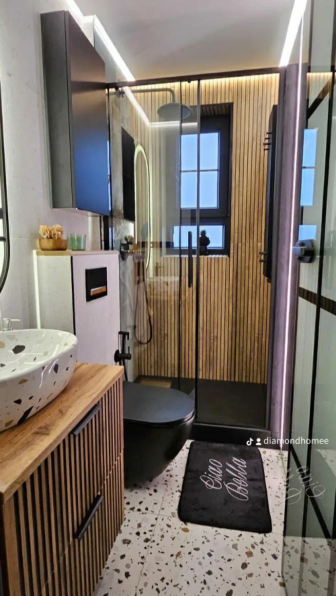 Nowoczesna łazienka z kabiną prysznicową typu walk-in, umywalką nablatową w kolorze lastryko, szafką z lamelami drewnianymi, czarną toaletą i dywanikiem z napisem 'Ciao Bella', podświetleniem LED...