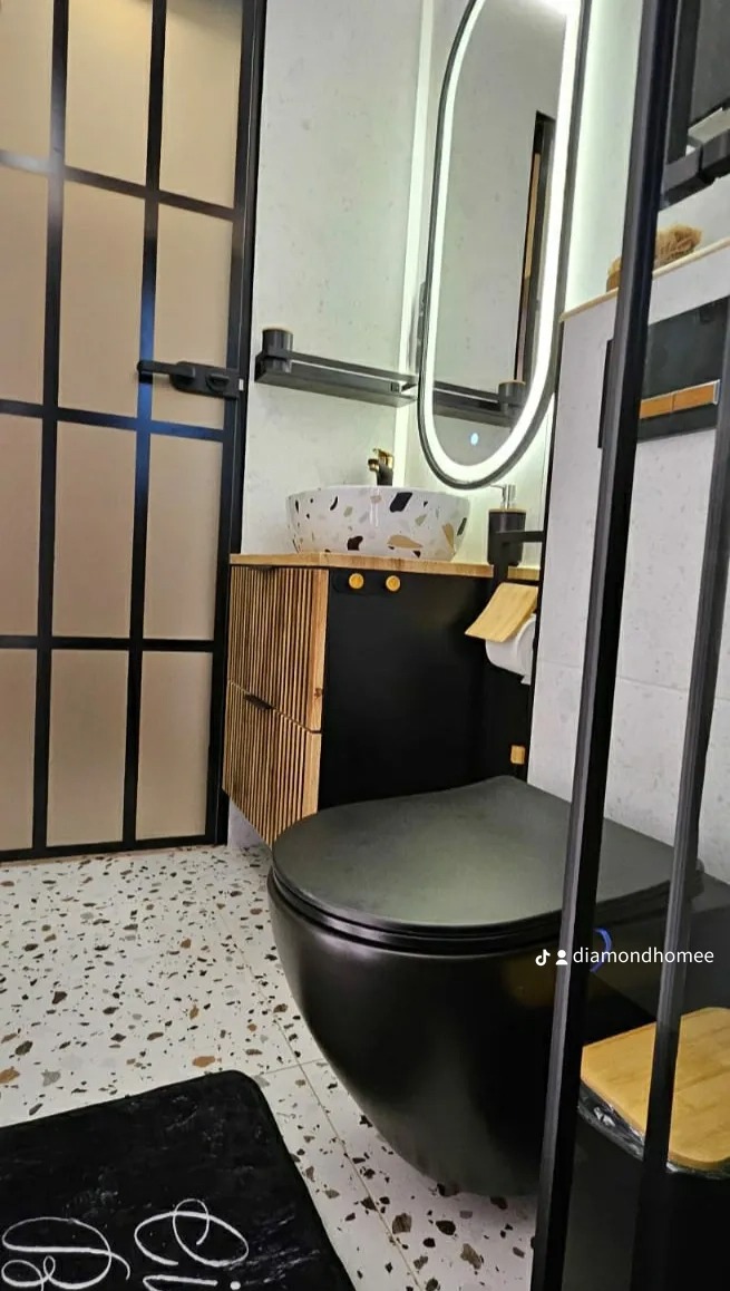 Nowoczesna łazienka z czarną miską WC wiszącą, umywalką nablatową w kolorze lastryko, szafką pod umywalką z lamelami i podświetlanym lustrem owalnym.