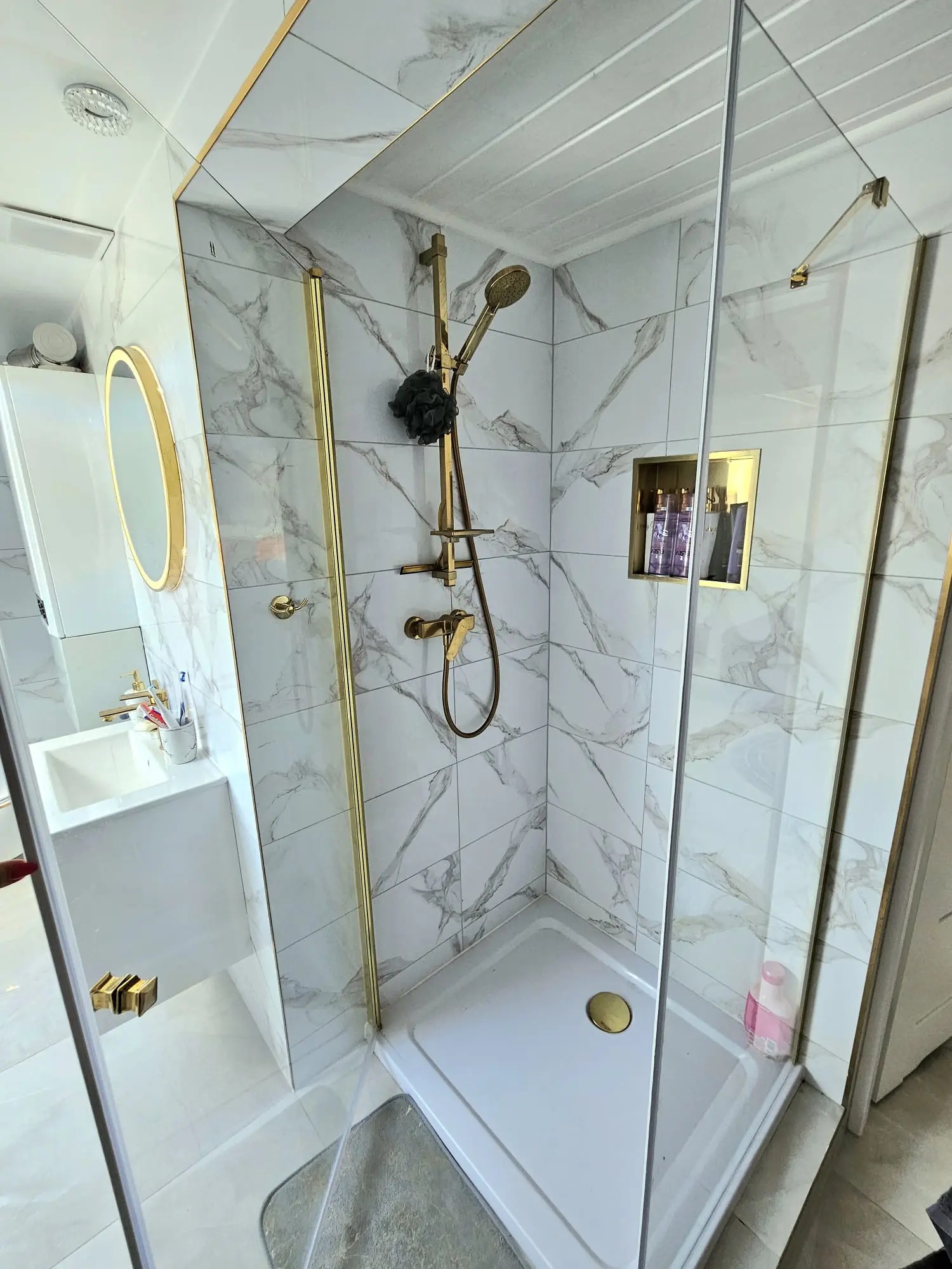 Nowoczesna łazienka z kabiną prysznicową typu walk-in, wykończona białymi płytkami imitującymi marmur z elementami w kolorze złotym, w tym armatura, rama kabiny i obramowanie lustra.