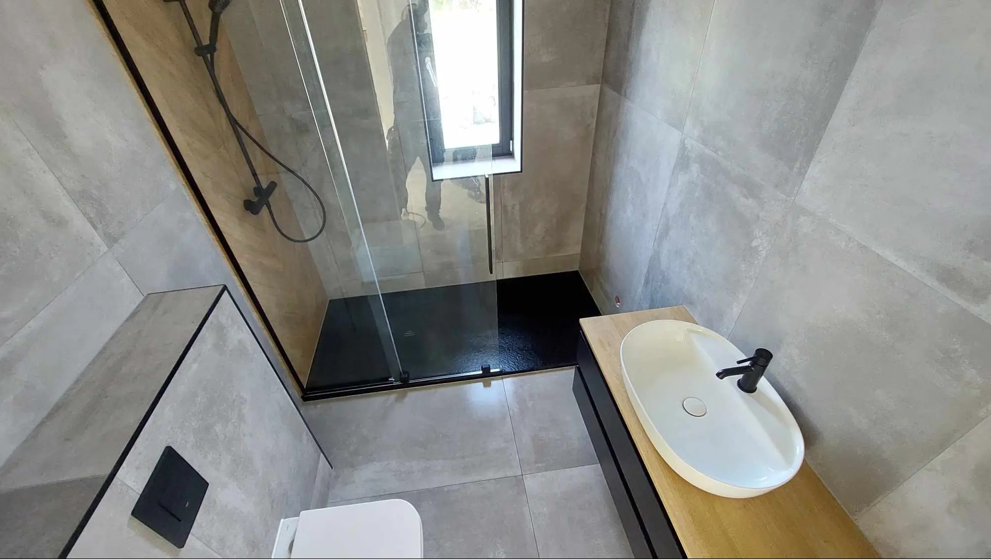 Nowoczesna łazienka z szarymi płytkami, czarną kabiną prysznicową typu walk-in z odpływem liniowym oraz owalną białą umywalką nablatową z czarną armaturą na drewnianym blacie.