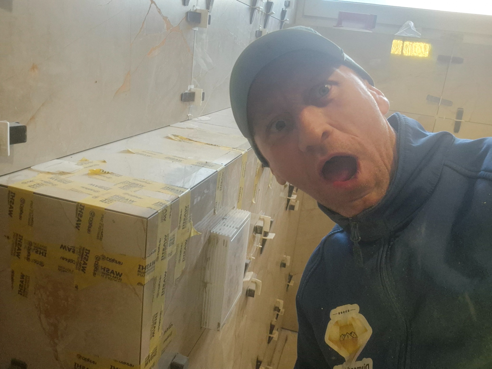 Mężczyzna z otwartymi ustami w bluzie i czapce na tle ściany z jasnych płytek, częściowo oklejonych taśmą, widoczne kliny poziomujące płytki.