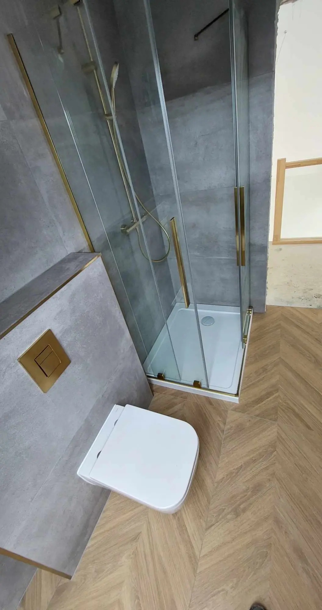Szklana kabina prysznicowa z złotymi wykończeniami, wisząca toaleta z przyciskiem w kolorze złota, szara płytka ścienna i podłoga z parkietu ułożonego w jodełkę.