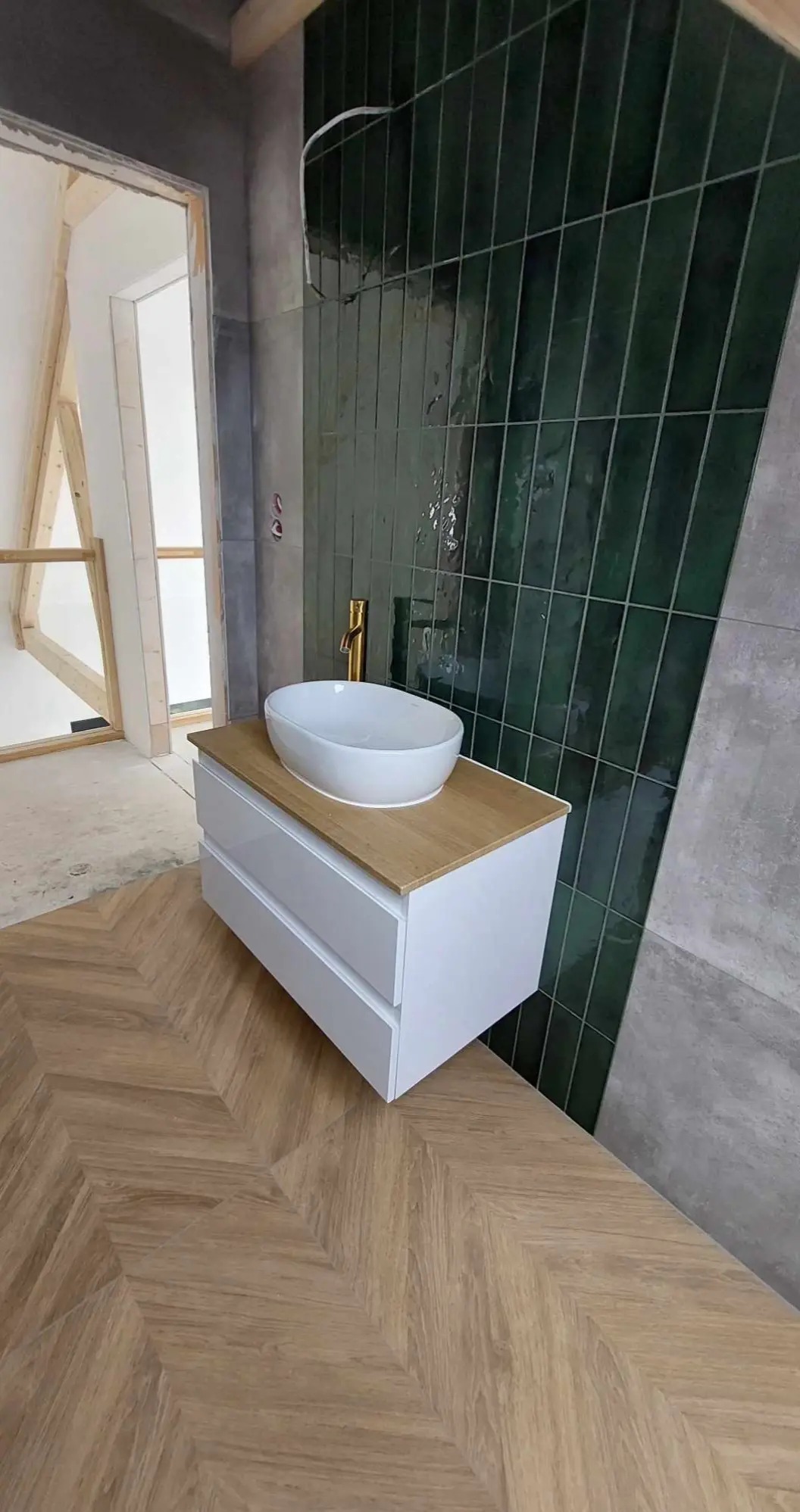 Umywalka nablatowa owalna z baterią w kolorze złotym, zamontowana na szafce z dwiema szufladami, na podłodze parkiet ułożony w jodełkę, w tle ściana wyłożona wąskimi, zielonymi płytkami.