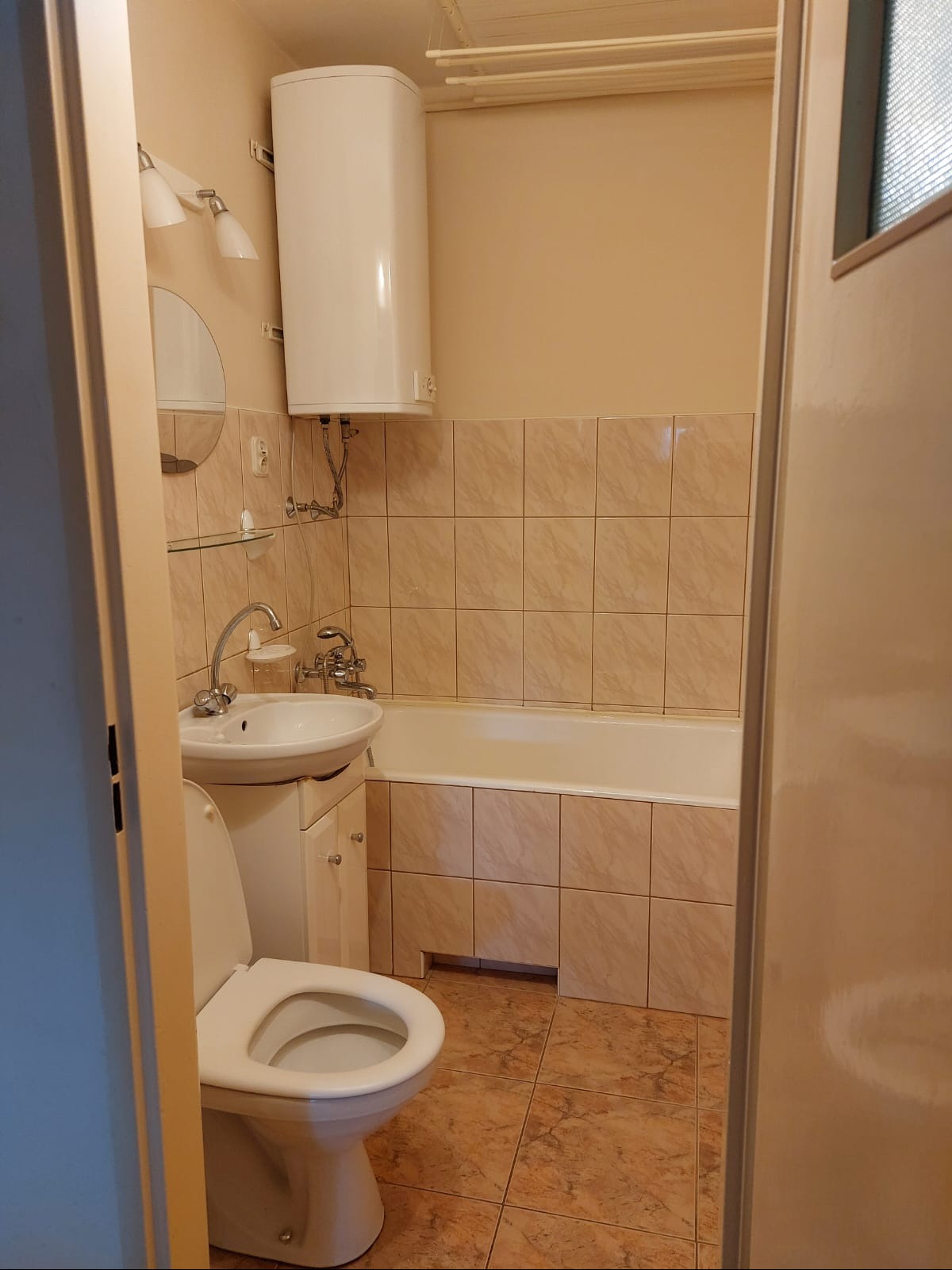 Ustawna łazienka w beżowych tonacjach z wanną obudowaną płytkami, umywalką z szafką i toaletą. Na ścianie bojler i lustro z oświetleniem. Widok zza uchylonych drzwi.