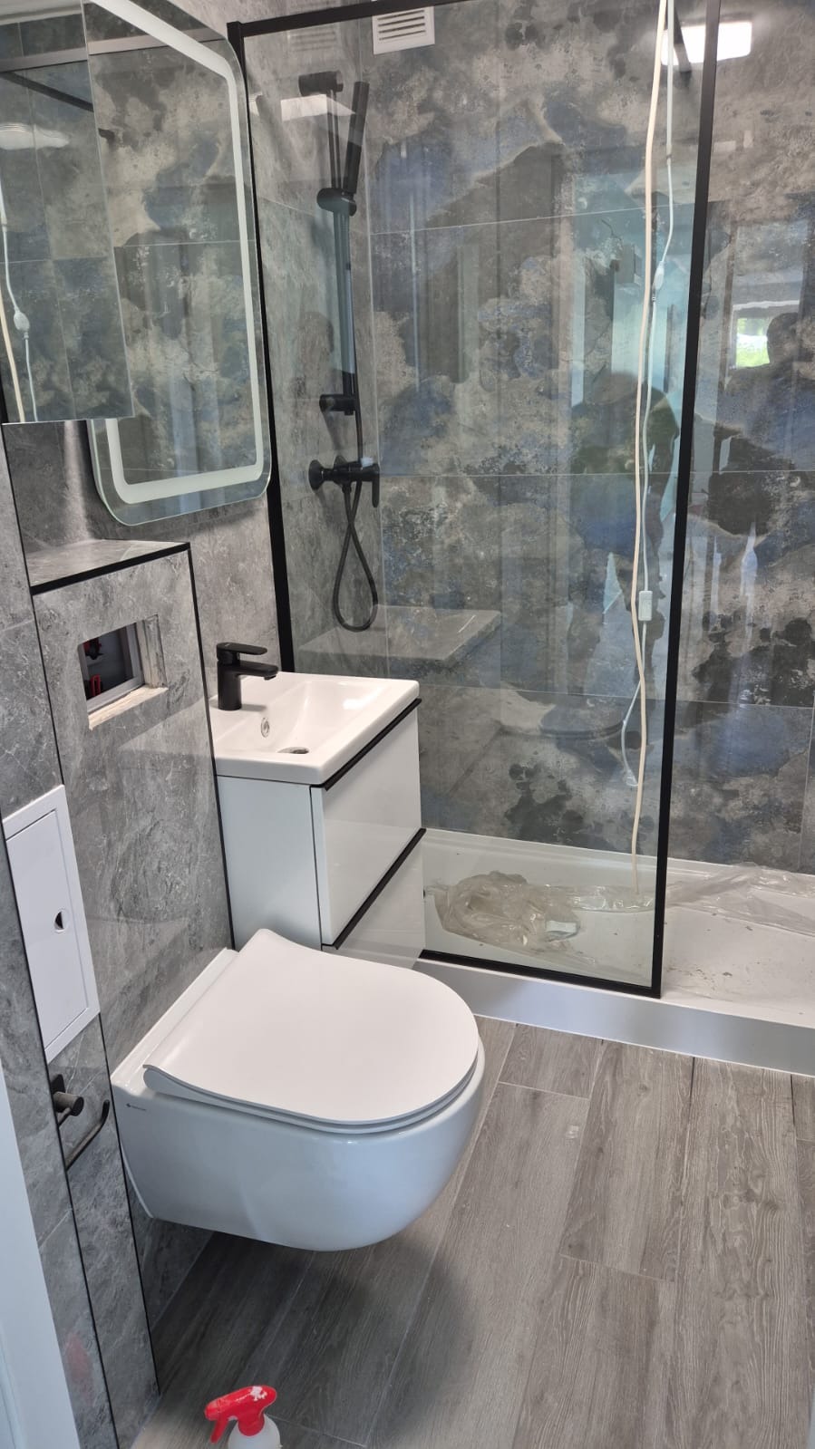 Nowoczesna łazienka po remoncie: wisząca toaleta, umywalka z szafką, kabina prysznicowa z czarnymi ramami, szare płytki imitujące kamień i drewnopodobna podłoga.