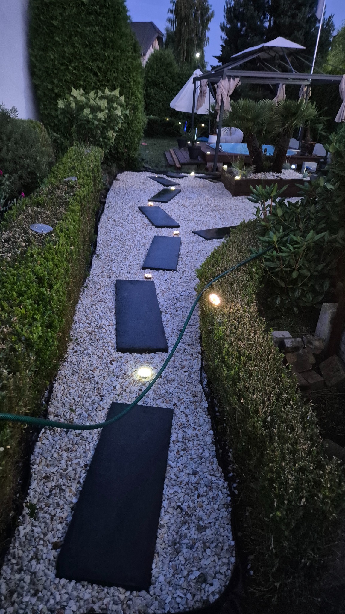 Oświetlona ścieżka z płyt i białego żwiru w ogrodzie, otoczona równo przyciętymi żywopłotami. W tle widoczny basen i altana.