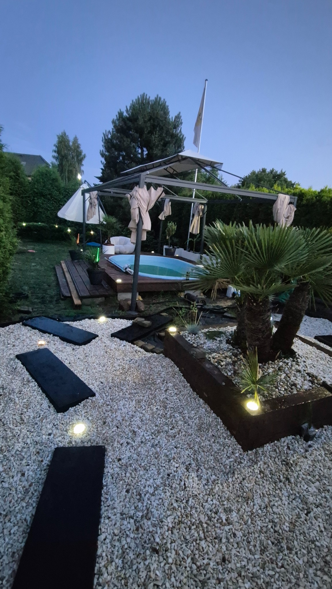 Ogród z basenem i palmą, oświetlony punktowymi lampami w kamiennym podłożu. Widoczna drewniana ścieżka i altana z zasłonami. Ujęcie z dołu, wieczorne oświetlenie.
