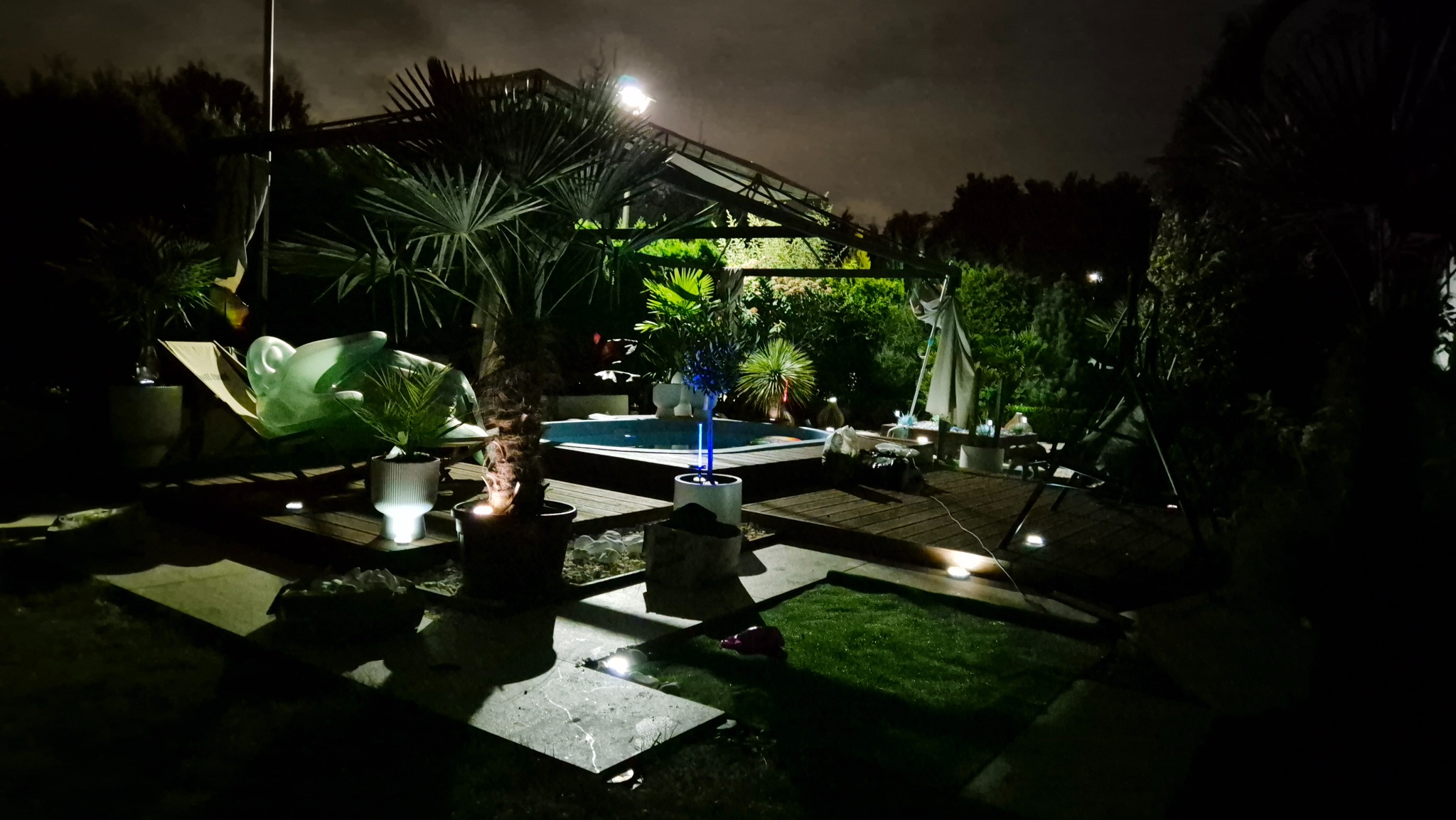 Nocne ujęcie podświetlonego ogrodu z basenem, palmami i drewnianym tarasem. Dekoracje świetlne i rzeźba dodają unikalnego charakteru.