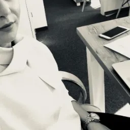 Kobieta w biurze, siedzi przy biurku z telefonem i laptopem. Na nadgarstku zegarek. Zdjęcie czarno-białe, z bliska. Częściowo widoczna twarz.