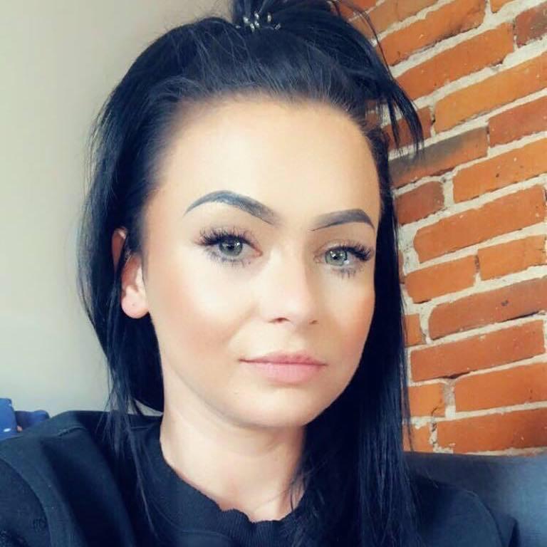 Portret kobiety o jasnych oczach i ciemnych włosach na tle ceglanej ściany. Uwagę zwracają długie rzęsy i subtelny makijaż. Ujęcie od przodu, skupione na twarzy.