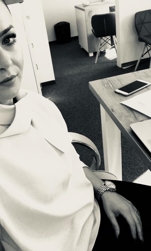 Kobieta w biurze, siedzi przy biurku z telefonem i laptopem. Na nadgarstku zegarek. Zdjęcie czarno-białe, z bliska. Częściowo widoczna twarz.