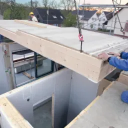 Montaż prefabrykowanego elementu stropu na budowie domu; robotnik w niebieskim kombinezonie i żółtym kasku układa element, w tle widoczny inny pracownik i zabudowa jednorodzinna.