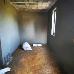 Wnętrze pomieszczenia w trakcie prac tynkarskich maszynowych: świeżo otynkowane ściany w odcieniach szarości, widoczne worki z materiałami budowlanymi na ziemi i surowy sufit z betonowych bloczków.