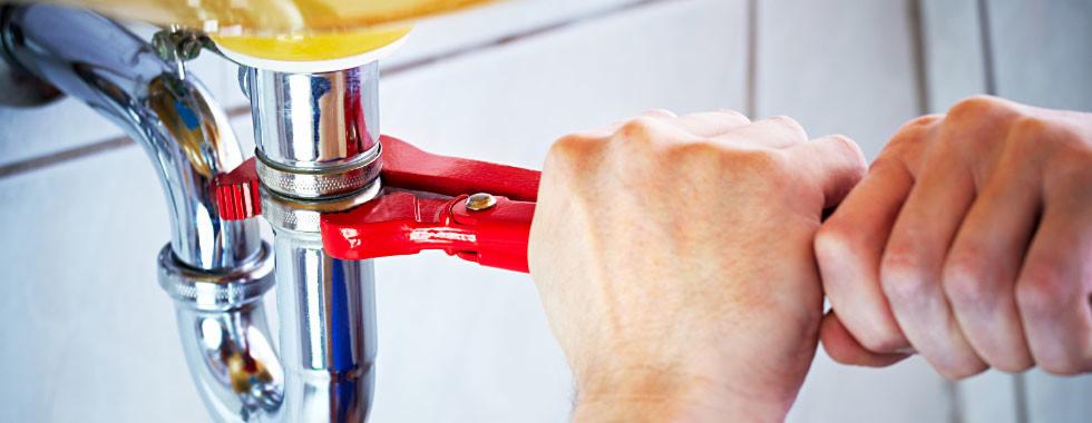 Instalacja chromowanej rury odpływowej pod umywalką, dokręcana czerwoną żabką przez hydraulika, widoczne białe płytki w tle.