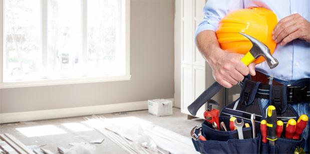 Mężczyzna trzymający kask i młotek, z pasem narzędziowym w pomieszczeniu z oknem i przygotowanym parkietem.