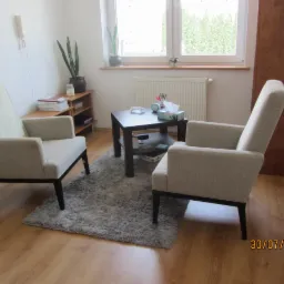 Jasny gabinet z dwoma fotelami, stolikiem kawowym i dywanem, przygotowany do sesji terapeutycznej. Na ścianie widoczny telefon. Zdjęcie z datą w prawym dolnym rogu.