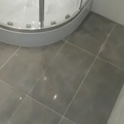 Szare, kwadratowe płytki podłogowe w łazience, ułożone przy narożnej kabinie prysznicowej z widocznymi kroplami wody na szybie i jasnymi fugami.