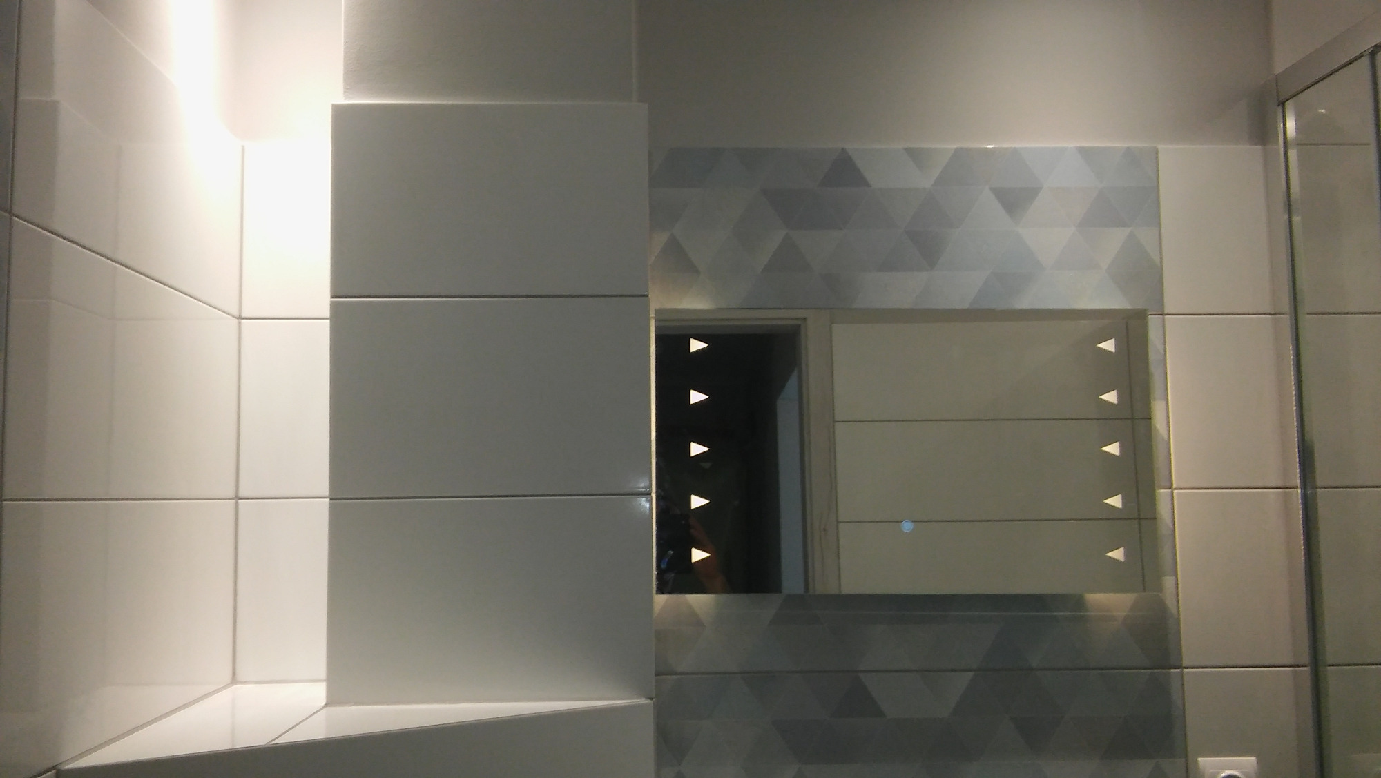 Nowoczesna łazienka z białymi płytkami, lustrem z podświetleniem LED i dekoracyjną ścianą z geometrycznym wzorem w odcieniach szarości.