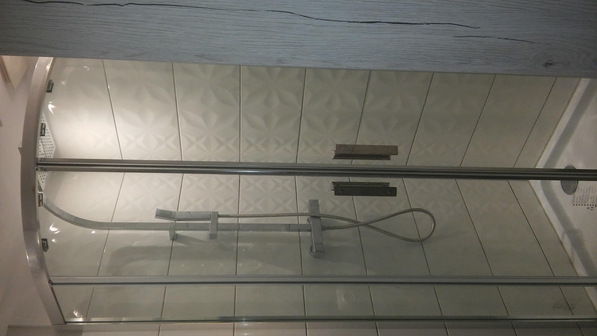 Szklana kabina prysznicowa z zamontowaną deszczownicą na tle ściany wyłożonej jasnymi, trójwymiarowymi płytkami ceramicznymi o wzorze geometrycznym.