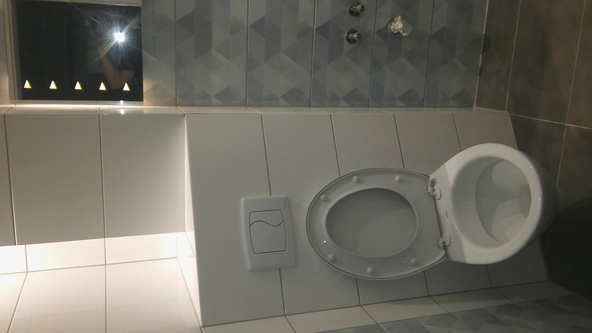 Wykończona toaleta z białą muszlą klozetową z otwartą deską, przycisk spłuczki na ścianie wyłożonej białymi i szarymi płytkami z geometrycznym wzorem oraz podświetlane lustro z odbiciem osoby.