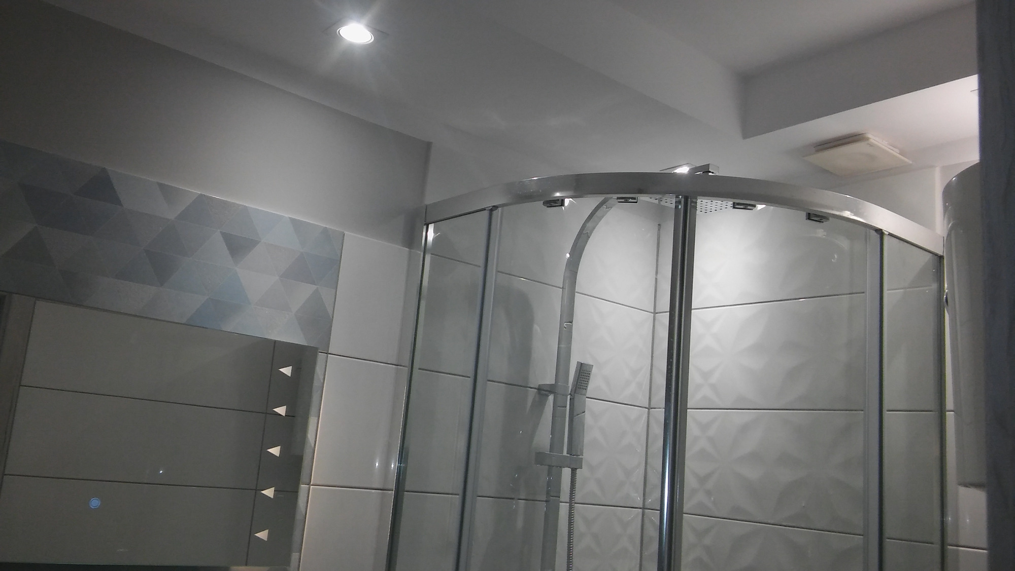 Szklana kabina prysznicowa z chromowanymi profilami na tle białych płytek z geometrycznym wzorem, oświetlona punktowym światłem sufitowym.