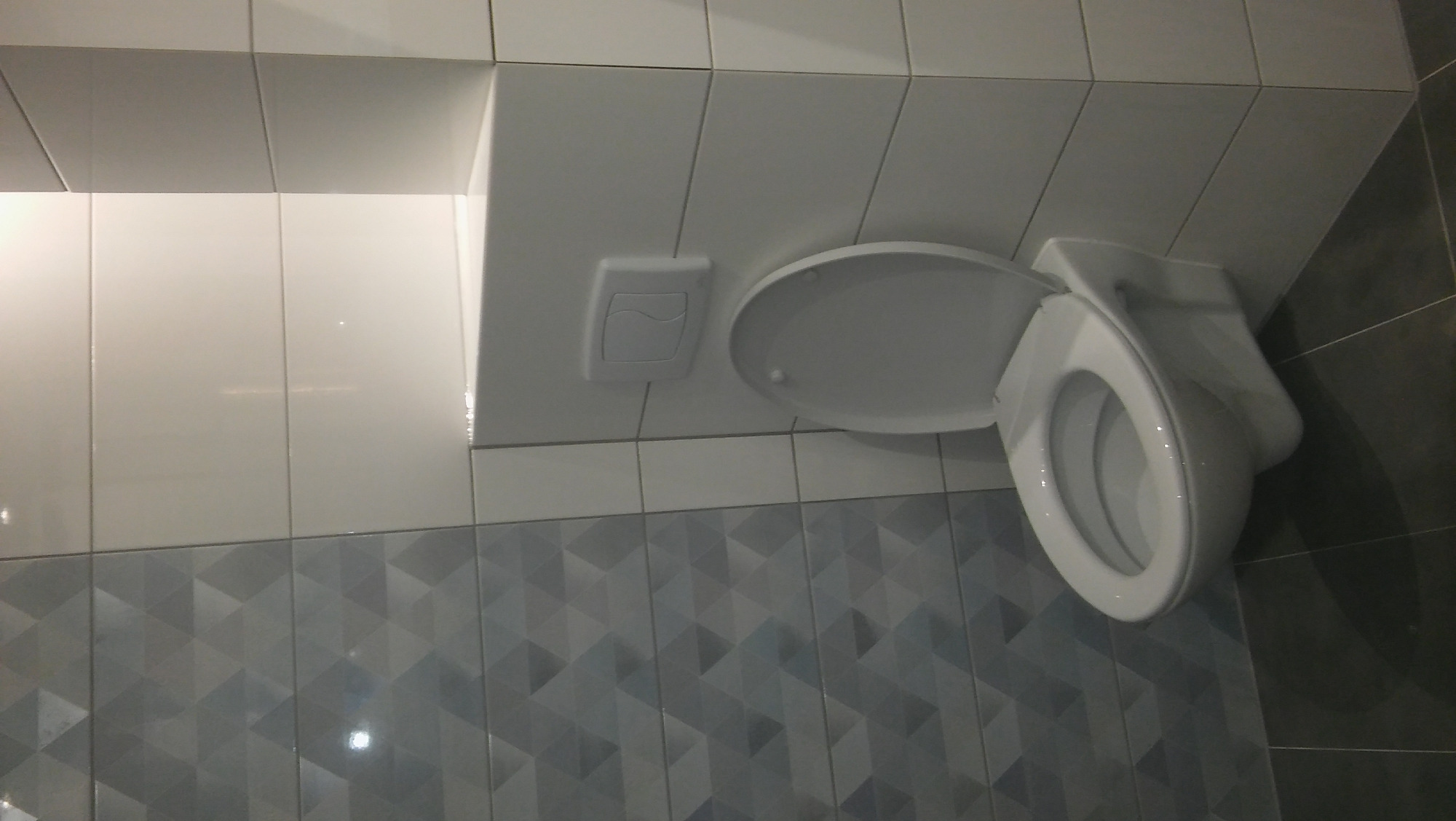 Podwieszana toaleta z otwartą klapą, białe płytki na ścianie i szare płytki z geometrycznym wzorem poniżej, widoczny przycisk spłuczki.