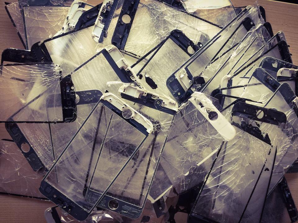 Stos potłuczonych ekranów telefonów komórkowych, widoczne liczne pęknięcia i uszkodzenia, niektóre zdemontowane z obudów, leżące na drewnianym podłożu.