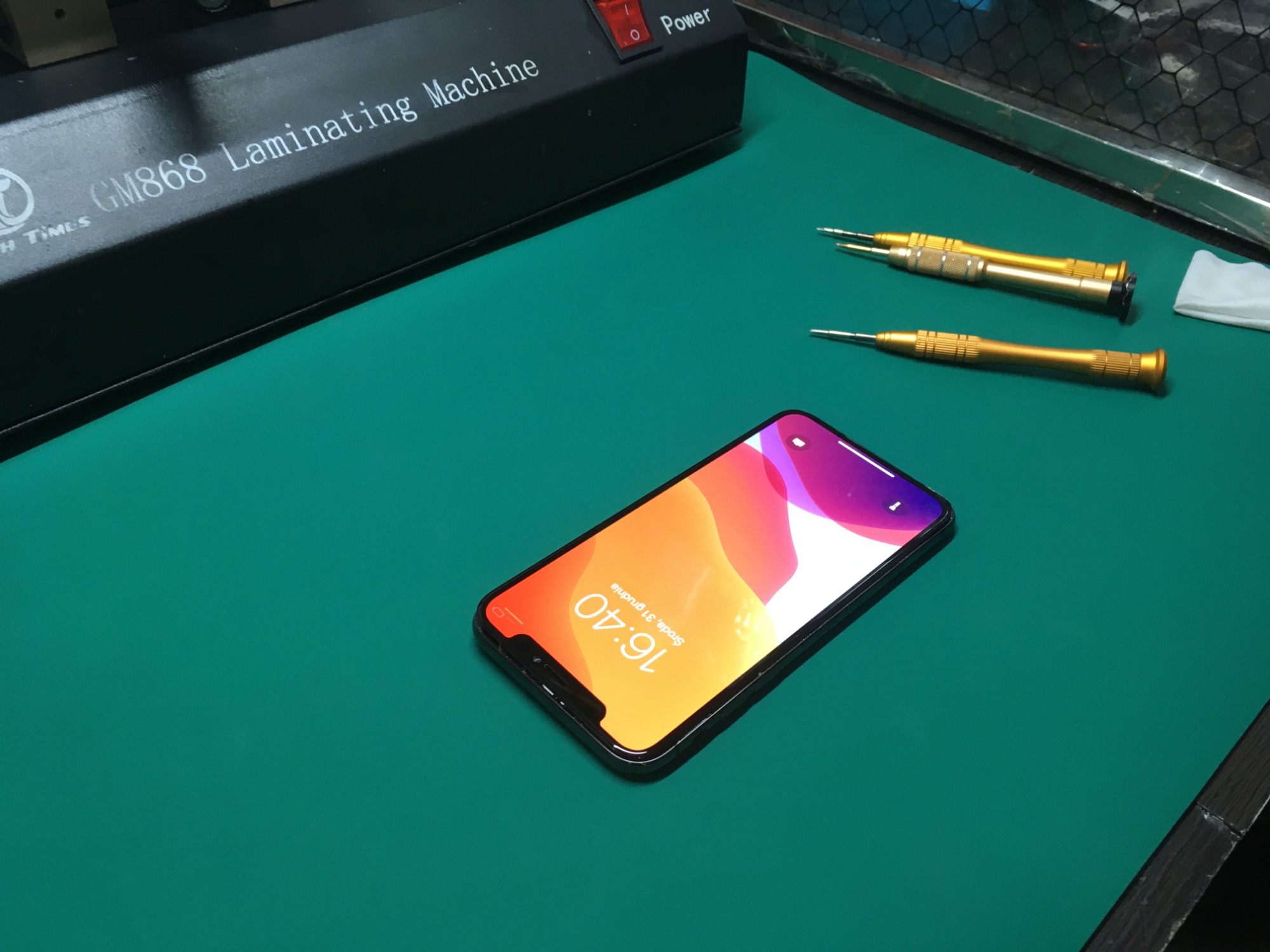 Smartfon z włączonym ekranem leżący na zielonej macie serwisowej obok złotych precyzyjnych śrubokrętów i laminatora GM868