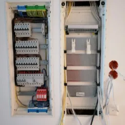 Dwie szafki elektryczne zamontowane na białej ścianie, widoczne liczne przewody, bezpieczniki i listwy zaciskowe wewnątrz szafek, po prawej stronie wystające przewody z puszki elektrycznej.