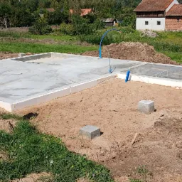 Wylana płyta fundamentowa z widocznymi przyłączami instalacji sanitarnych, na piaszczystym podłożu z elementami szalunków i narzędzi budowlanych w tle wiejski krajobraz.
