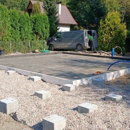 Woodwork - Przygotowany teren pod budowę, z widocznymi betonowymi bloczkami na żwirowym podłożu, izolacją i pracownikiem przy samochodzie dostawczym w tle.