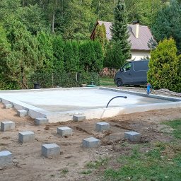 Woodwork - Wylany fundament pod budowę domu z widocznymi betonowymi słupkami na obrzeżach, niebieska przenośna toaleta Toalet.pl z boku, w tle zielony ogród i szary samochód dostawczy.