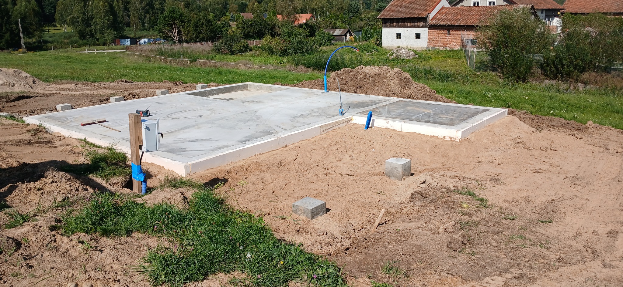 Wylana płyta fundamentowa z widocznymi przyłączami instalacji sanitarnych, na piaszczystym podłożu z elementami szalunków i narzędzi budowlanych w tle wiejski krajobraz.