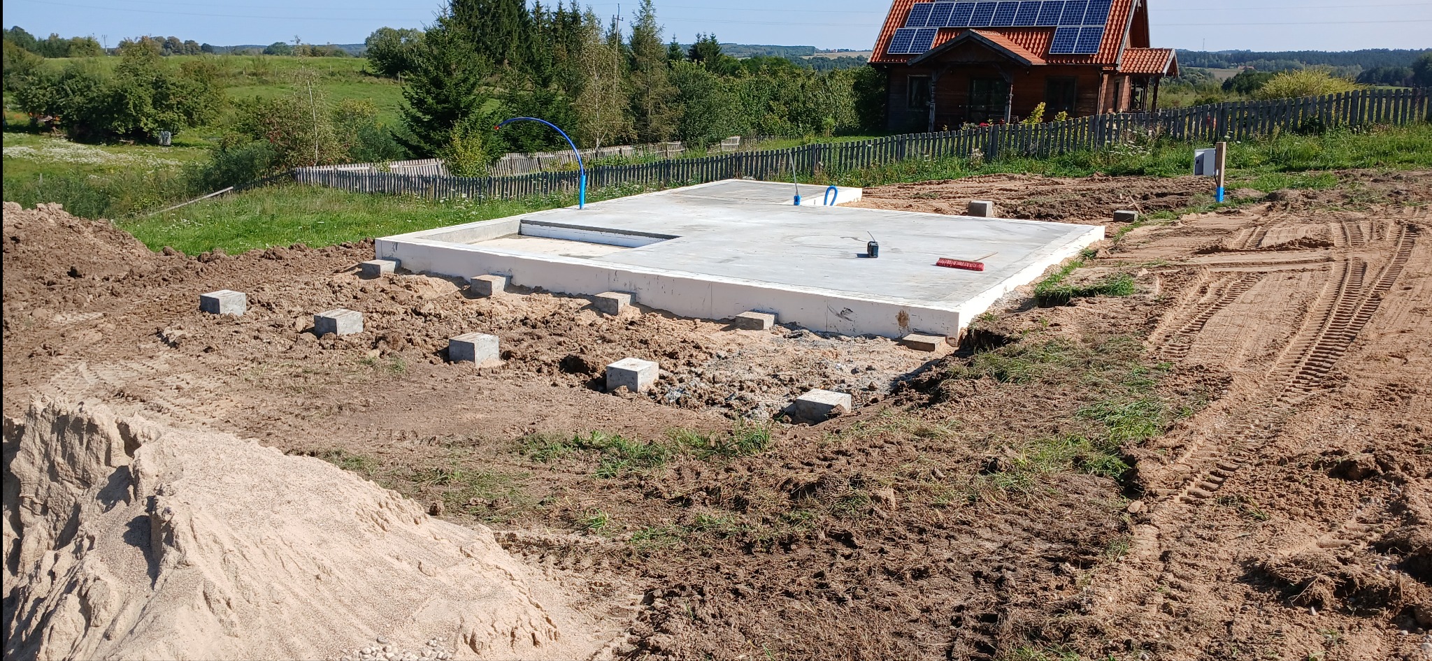 Wylany fundament pod budynek z widocznymi przyłączami instalacji, bloczkami betonowymi pod przyszłe ściany i śladami opon maszyn budowlanych na ziemi.