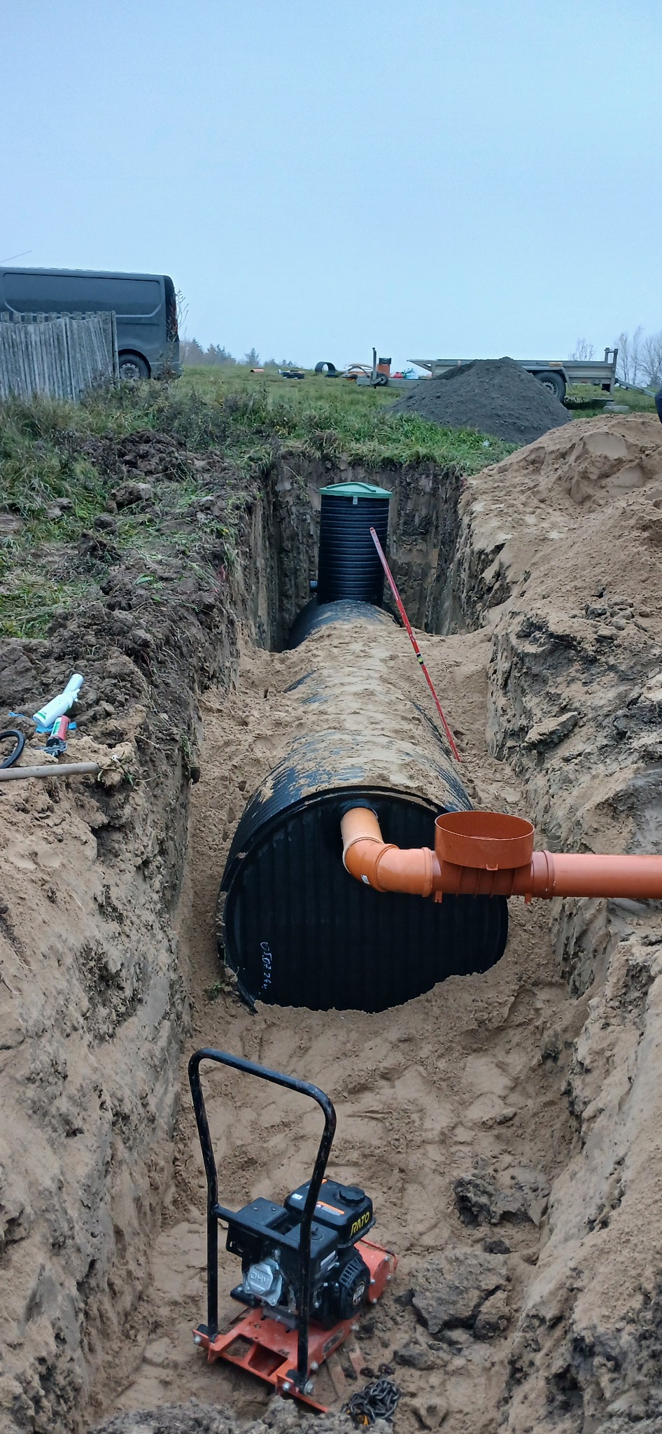 Czarny zbiornik na ścieki z podłączonymi pomarańczowymi rurami w głębokim wykopie, ubijarka spalinowa RATO na dnie wykopu, w tle widoczny zielony trawnik, szara furgonetka i sterta ziemi.
