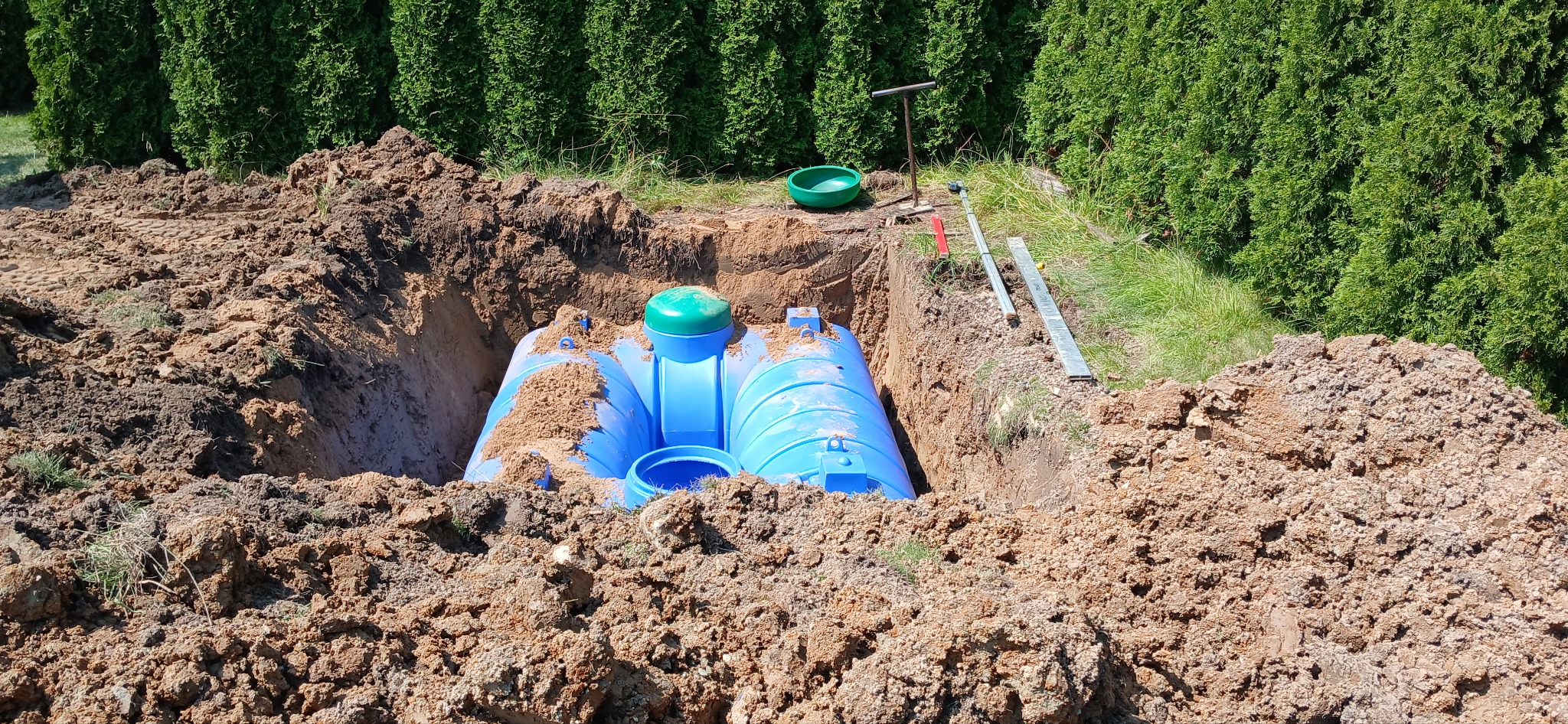 Niebieski zbiornik na ścieki z zieloną pokrywą, częściowo zasypany ziemią w wykopie na tle zielonych krzewów, widoczne narzędzia pomiarowe.